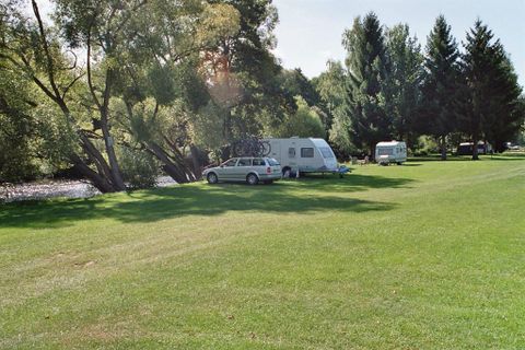 Ein ruhiger Campingplatz am Wasser mit grünen Wiesen. Zwei Wohnmobile stehen unter schattigen Bäumen.