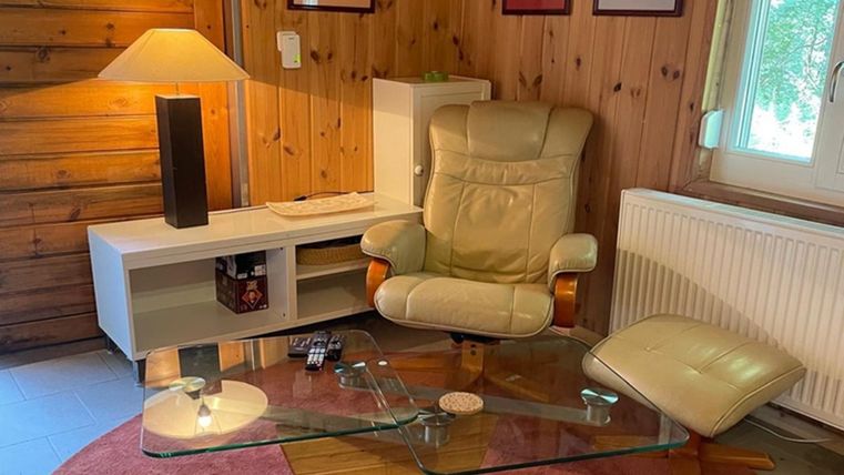 Ein gemütlicher Wohnbereich mit einem bequemen Stuhl und einem Glastisch. Die Wände sind aus Holz und es gibt einen warmen Lichtschein von einer Lampe.