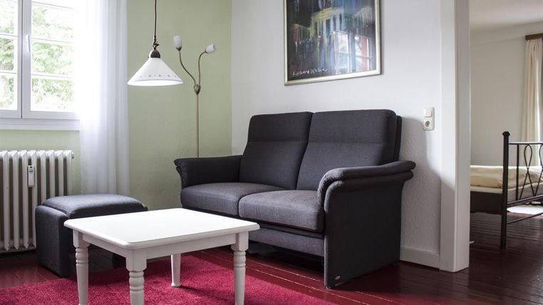 Ein gemütliches Wohnzimmer mit einer grauen Couch und einem kleinen Tisch. Der Raum hat helle Wände und ein rotes Teppich.