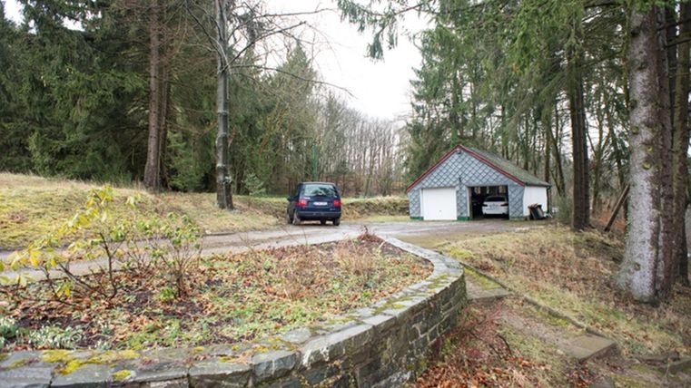 Eine ruhige Landschaft mit einem Auto, das vor einer Garage steht. Umgeben von Bäumen und einer gut gepflegten Gartenlandschaft.