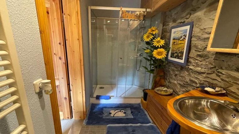 Ein modernes Badezimmer mit einer gläsernen Duschkabine und einem Waschbecken aus Holz. Bunte Badteppiche und eine Blumendekoration sorgen für eine einladende Atmosphäre.