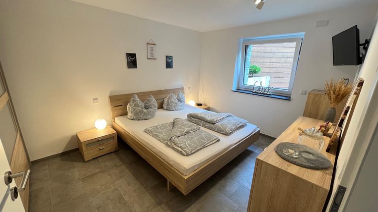 Ein modernes Schlafzimmer mit einem Doppelbett, Nachttischen und einem Fernseher. Der Raum hat helle Wände und ein großes Fenster, das Tageslicht hereinlässt.