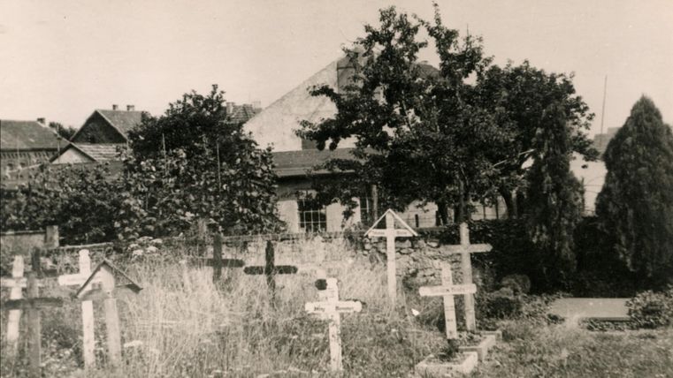 Ein alter Friedhof mit mehreren Holzkreuzen und hohem Gras. Im Hintergrund sind Gebäude und Bäume sichtbar.