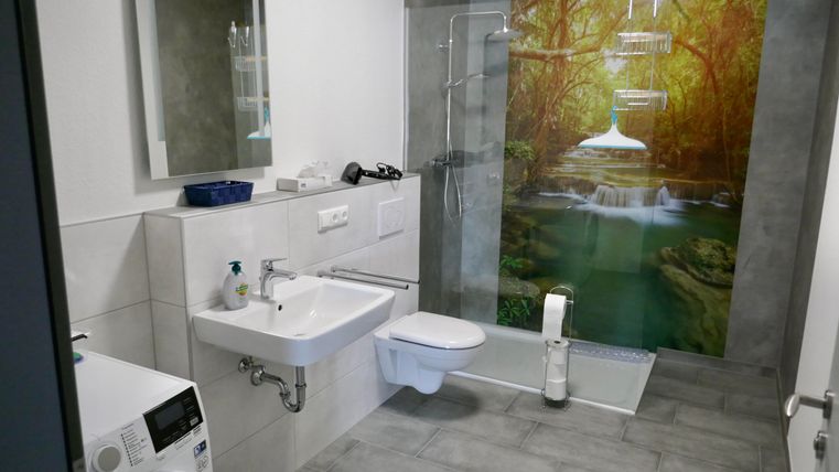 Ein modernes Badezimmer mit einer Dusche und einem Waschbecken. Die Wand ist mit einem Motiv von einem Wasserfall und einer grünen Landschaft gestaltet.