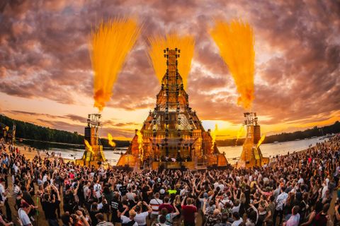 Eine große Menschenmenge sieht sich ein spektakuläres Event bei Sonnenuntergang an. Im Hintergrund steht eine beeindruckende Konstruktion mit bunten Feuerwerkseffekten.