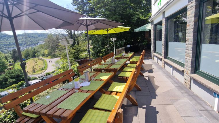 Eine einladende Terrasse mit Holztischen und gelben Sonnenschirmen. Im Hintergrund sind grüne Hügel zu sehen.