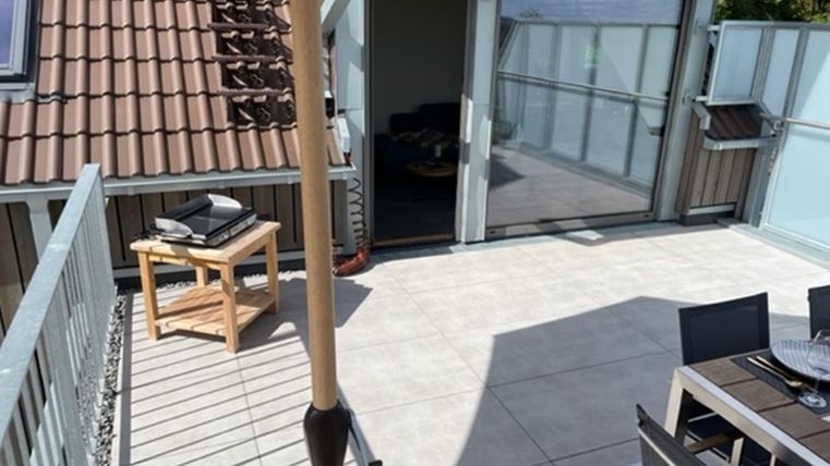 Eine Terrasse mit modernen Möbeln und einem Sonnenschirm. Im Hintergrund ist ein Glas-Schiebetür zu sehen, die ins Innere führt.