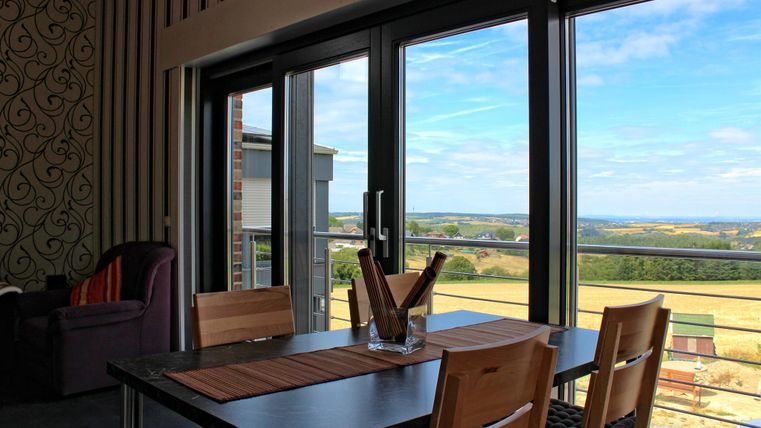Ein heller Raum mit einem Esstisch, Stühlen und großen Fenstern, die eine schöne Aussicht auf die Landschaft bieten. Der Raum hat eine moderne Einrichtung mit einer gemütlichen Ecke.