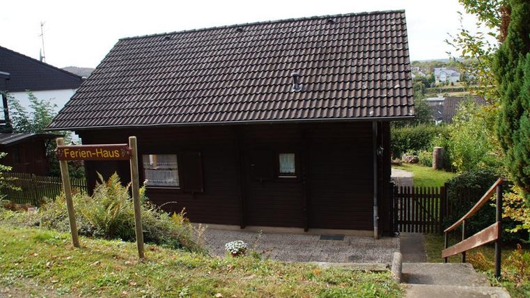 Ein rustikales Ferienhaus mit brauner Holzfassade. Es steht in einer grünen Umgebung mit einem kleinen Garten und einem Weg zur Haustür.