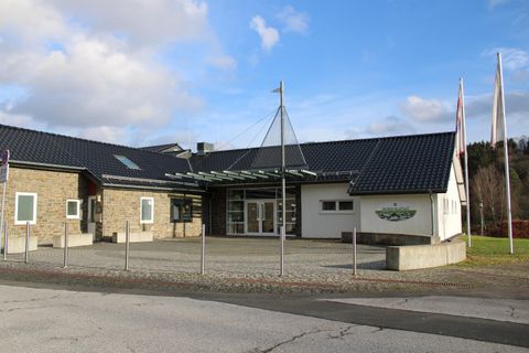 Ein modernes Gebäude mit einem Flachdach und großen Fenstern. Vor dem Eingang sind mehrere Flaggen und ein gepflasterter Bereich.