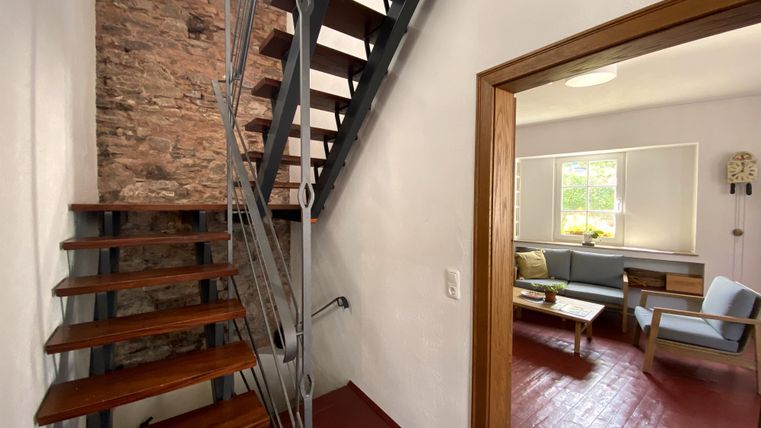 Ein moderner Flur mit einer Treppe aus Holz und Metall. Im Hintergrund ist ein gemütlicher Wohnbereich mit einem Sofa und einem Tisch sichtbar.