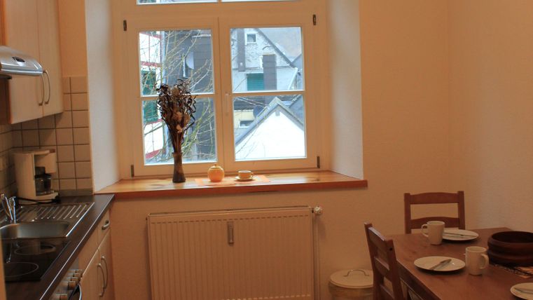 Eine helle Küche mit Holzfußboden und großen Fenstern. Ein Tisch mit Geschirr steht neben der Kochzeile.