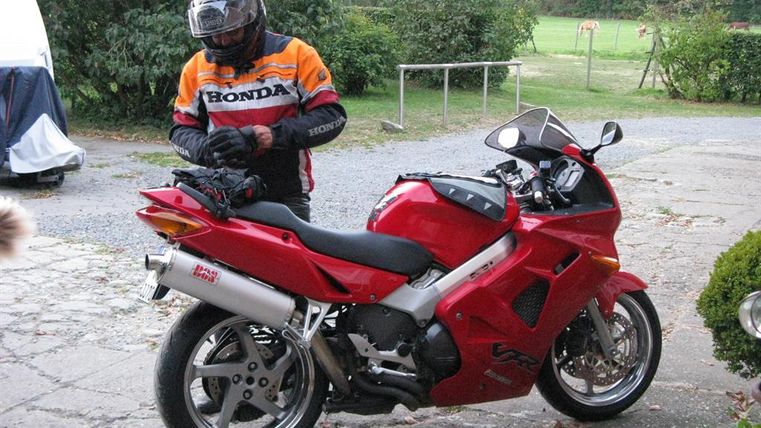 Ein Motorradfahrer in Schutzkleidung steht neben einem roten Motorrad. Die Umgebung ist grün und ländlich, mit einem klaren Himmel im Hintergrund.