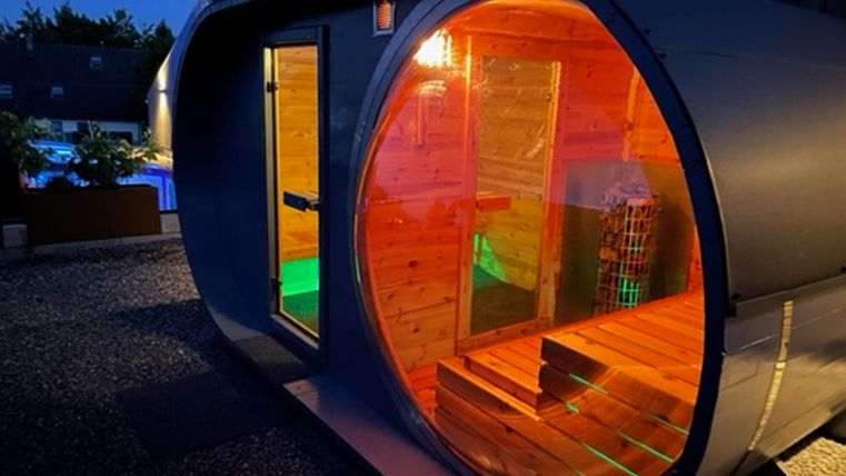Eine moderne Sauna in runder Form, erleuchtet mit bunten Lichtern. Sie steht in einem Garten bei Nacht und verfügt über eine Holzverkleidung.