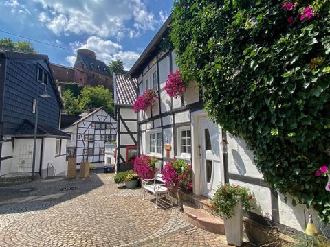 Ein malerischer Innenhof mit Fachwerkhäusern und bunten Blumen. Im Hintergrund sind grüne Bäume und ein Schloss sichtbar.