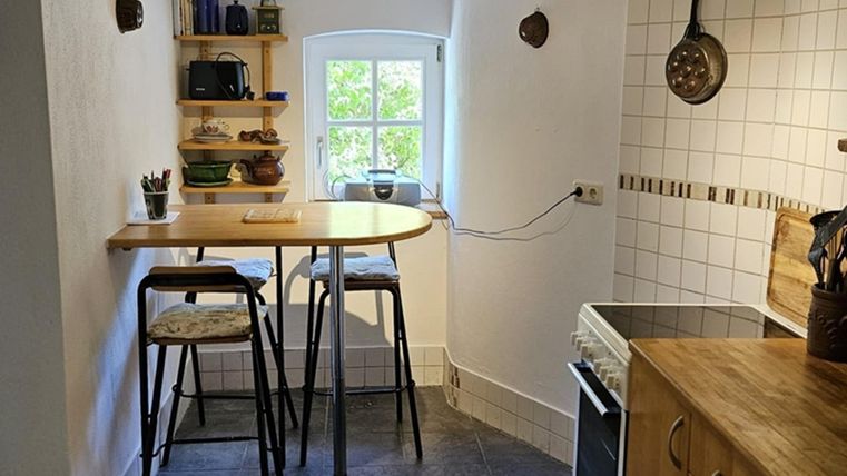 Eine gemütliche Küche mit einem kleinen Tisch und hochkantigen Stühlen. An der Wand befinden sich offene Regale mit Geschirr und Küchenutensilien.