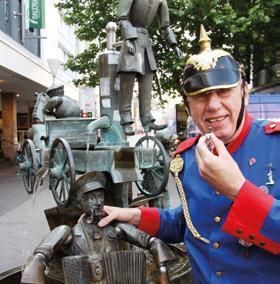 Ein Mann in einer historischen Uniform steht neben einer Brunnenfigur. Im Hintergrund sind weitere Figuren und ein Wagen zu sehen.