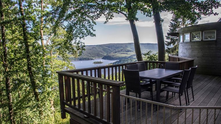 Eine Terrasse mit Holzmöbeln und Blick auf einen See, umgeben von Bäumen. Die Landschaft ist hügelig und strahlt Ruhe aus.