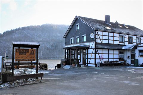 Ein malerisches Gasthaus mit Fachwerkarchitektur am Wasser. Im Hintergrund sind bewaldete Hügel zu sehen.