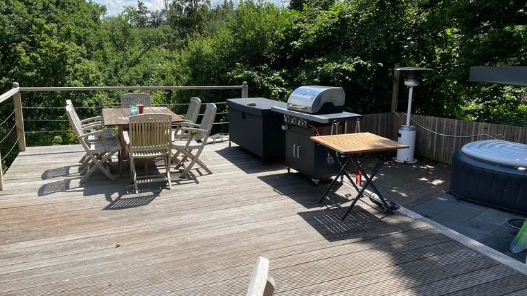 Eine gemütliche Terrasse mit einem Grill und einem Esstisch. Im Hintergrund sind viele Bäume und blauer Himmel sichtbar.
