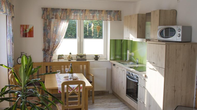 Eine helle Küche mit modernen Möbeln und einer großen Fensterfront. Der Tisch ist für vier Personen gedeckt und Pflanzen sorgen für eine angenehme Atmosphäre.