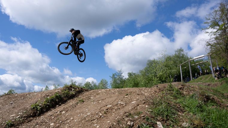 Ein Mountainbiker springt mit seinem Fahrrad über einen Hügel. Der Himmel ist blau mit einigen Wolken und die Umgebung ist grün.