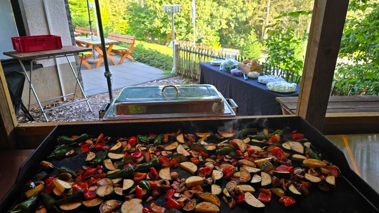 Ein Grill mit buntem Gemüse in einer schönen Gartenumgebung. Im Hintergrund sieht man einen gedeckten Tisch und eine grüne Landschaft.