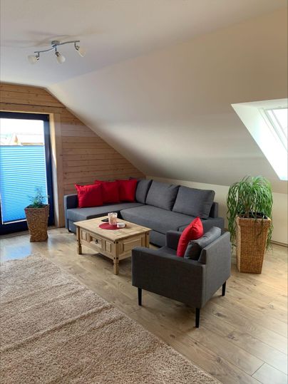 Ein gemütliches Wohnzimmer mit einem grauen Sofa und roten Kissen. Der Raum hat große Fenster und einen kleinen Tisch in der Mitte.