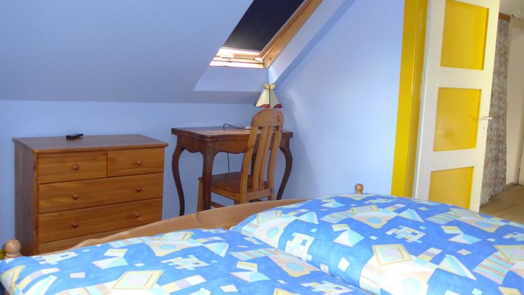 Ein gemütliches Schlafzimmer mit einem Bett, einer Kommode und einem Schreibtisch. Die Wände sind hellblau und eine Tür in Gelb fügt einen Farbtupfer hinzu.