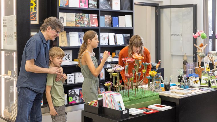 Eine Familie besucht einen Geschäft mit bunten Kunsthandwerken und Büchern. Die Kinder zeigen Interesse an den Produkten am Verkaufstresen.