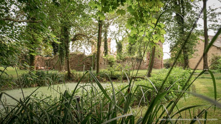 Ein ruhiger Garten mit üppigem Grün und Bäumen. Im Hintergrund sind alte Mauern sichtbar.