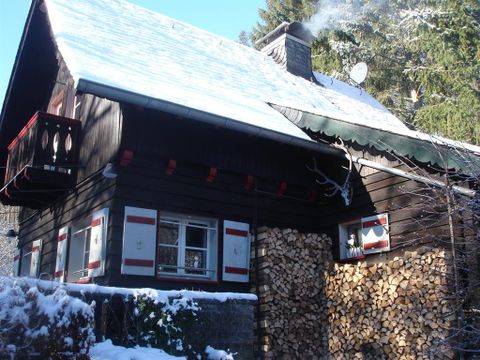 Ein gemütliches Holzhaus mit einem schneebedeckten Dach und rauchendem Schornstein. Neben dem Haus liegt ein Stapel Holz für den Winter.