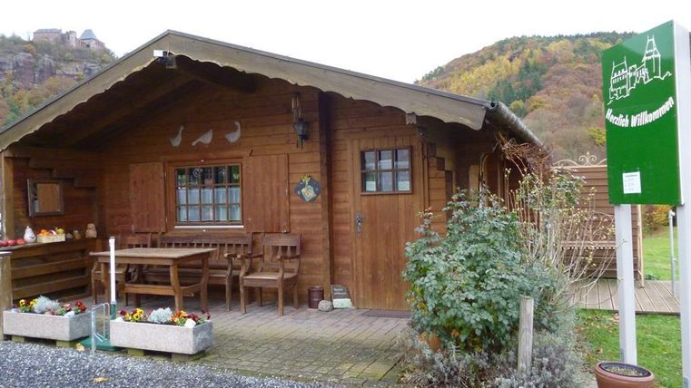 Ein gemütliches Holzhaus mit einer Terrasse und blühenden Pflanzen. Die Umgebung ist von sanften Hügeln und herbstlichen Farben geprägt.