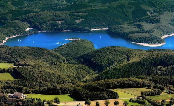 Eine malerische Landschaft mit einem blauen See, umgeben von grünen Hügeln und Wäldern. Segelboote gleiten ruhig über das Wasser.