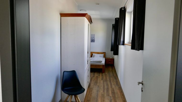 Ein einfaches Zimmer mit einem Bett, einem Kleiderschrank und einem Stuhl. Das Zimmer hat helle Wände und Holzfußboden.