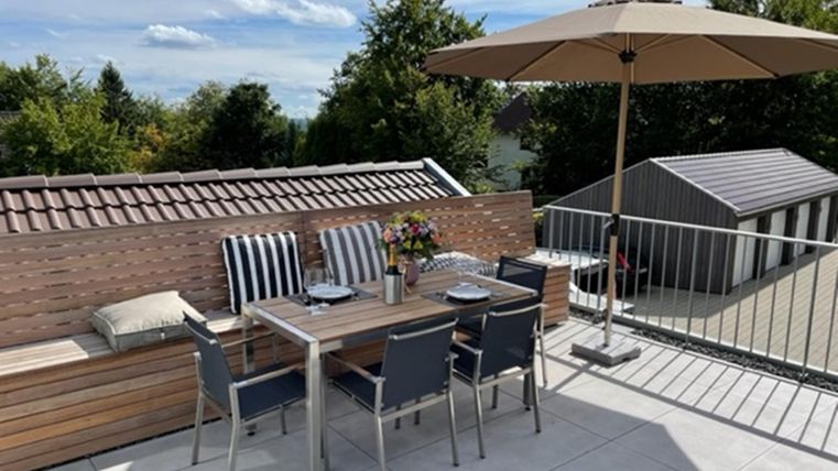 Eine gemütliche Terrasse mit einem Tisch und Stühlen. Ein Sonnenschirm bietet Schatten und Blumen dekorieren den Tisch.
