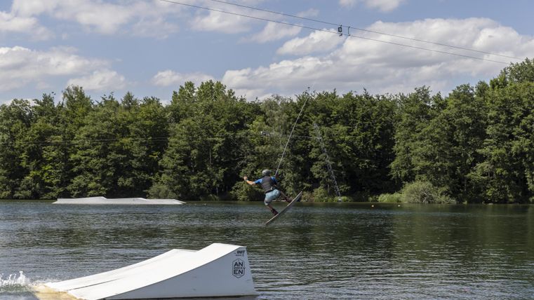Ein Wakeboarder springt über ein Hindernis auf einem ruhigen Gewässer. Im Hintergrund sind Bäume und ein blauer Himmel zu sehen.