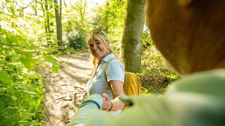Eine Frau lächelt auf einem sonnigen Waldweg und hält die Hand eines anderen. Die Umgebung ist grün und voller Bäume.