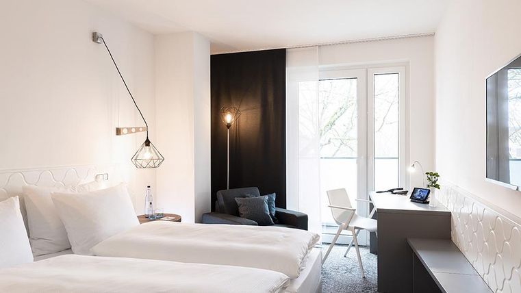 Ein modernes Hotelzimmer mit zwei Betten und einem gemütlichen Sitzbereich. Große Fenster sorgen für viel Tageslicht und eine freundliche Atmosphäre.
