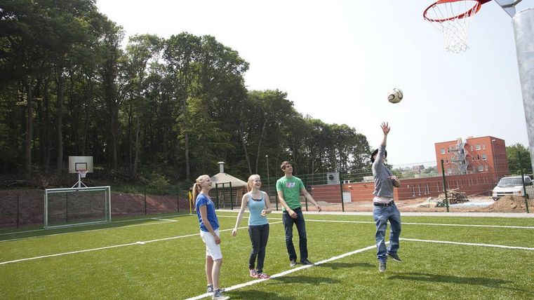 Eine Gruppe von jungen Menschen spielt Basketball auf einem Sportplatz mit Kunstrasen. Im Hintergrund sind Basketballkörbe und Bäume sichtbar.