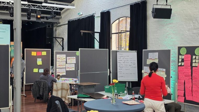 Ein kreativer Arbeitsraum mit verschiedenen Ständen und farbigen Post-its. Menschen sind damit beschäftigt, Ideen zu entwickeln und zu diskutieren.