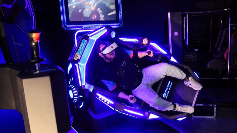 Eine Person sitzt in einem modernen VR-Simulator mit leuchtenden Lichtern. Auf dem Bildschirm sind interaktive Elemente sichtbar, die ein immersives Erlebnis zeigen.