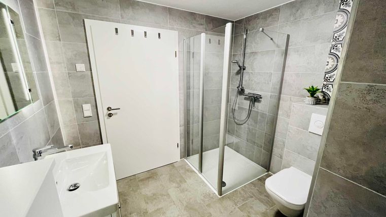 Ein modernes Badezimmer mit grauen Fliesen. Es verfügt über eine Dusche, ein Waschbecken und eine Toilette.