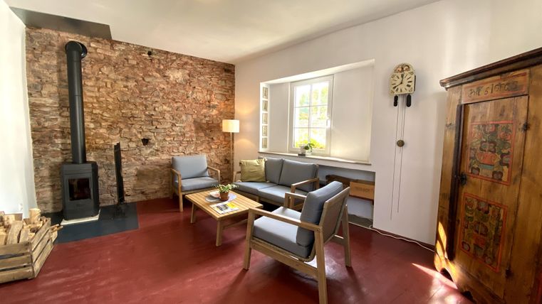 Ein gemütliches Wohnzimmer mit einem Holzkamin, blauen Sesseln und einem rustikalen Kasten. Die Wände sind teilweise mit Steinen verkleidet und es gibt viel natürliches Licht.