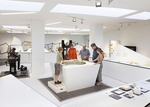 Eine moderne Ausstellung mit mehreren Besuchern, die an einem interaktiven Tisch stehen. Im Hintergrund sind einige Ausstellungsobjekte und Geräte zu sehen.