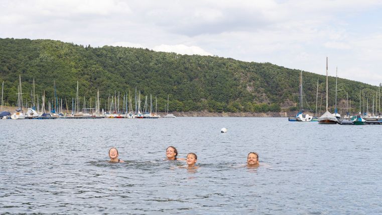 Vier Personen schwimmen im klaren Wasser eines Hafens. Im Hintergrund sind Boote und eine grüne Hügelkette zu sehen.
