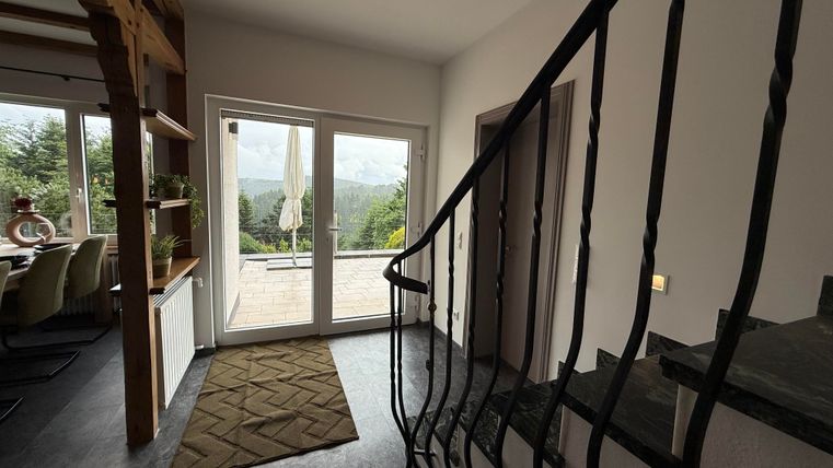 Ein heller Eingangsbereich mit einer geschwungenen Treppe und großen Fenstern. Im Hintergrund ist eine Terrasse mit Blick in die Natur sichtbar.
