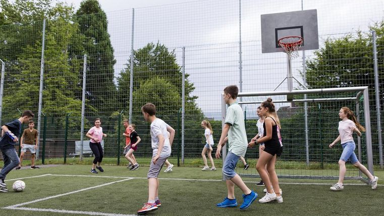 Eine Gruppe von Kindern spielt Fußball auf einem Sportplatz. Im Hintergrund ist ein Basketballkorb zu sehen.