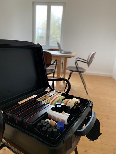 Ein offener Werkzeugkoffer mit verschiedenen Materialien steht auf einem Stuhl. Im Hintergrund sind ein Tisch und Stühle in einem hellen Raum zu sehen.