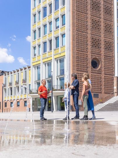 Eine Gruppe von vier Personen steht vor einem modernen Gebäude an einem sonnigen Tag. Der Platz ist nass, vermutlich von einem Springbrunnen.